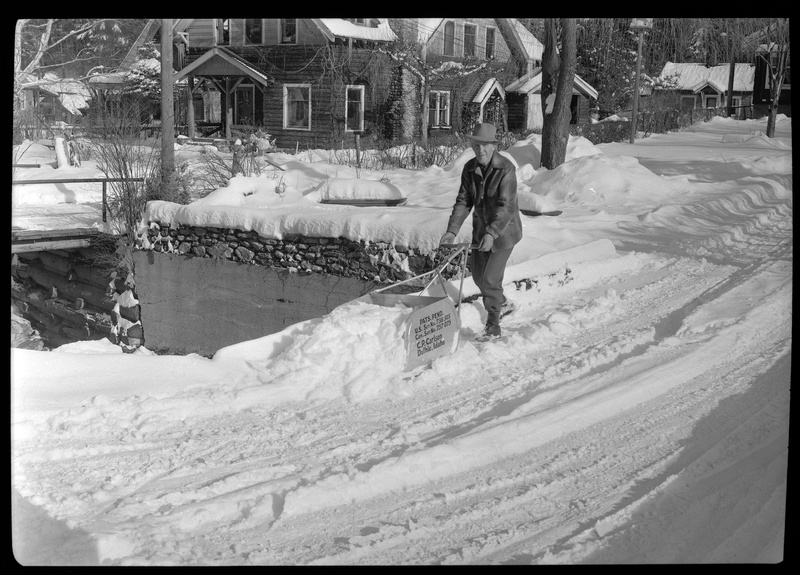 Photo of a man using a C. P. Carlson snow shovel to remove snow from the sidewalk. The shovel requires two hands to push it and holds more snow than the average snow shovel. There is a house behind the man. The side of the shovel reads: "Pats. Pend.; U.S. Ser. No. 738505; Can. Ser. No. 757075; C. P. Carlson; Duthie Idaho."