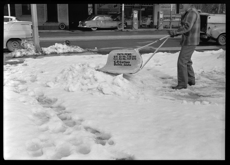 Photo of a man using a C. P. Carlson snow shovel to remove snow from the sidewalk. The shovel requires two hands to push it and holds more snow than the average snow shovel. The side of the shovel reads: "Pats. Pend.; U.S. Ser. No. 738505; Can. Ser. No. 757075; C. P. Carlson; Duthie Idaho." A road with cars on it can be seen behind the man.