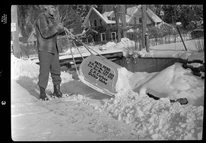 Photo of a man using a C. P. Carlson snow shovel to dump snow from the road. The shovel requires two hands to push it and holds more snow than the average snow shovel. There is a house behind the man. The side of the shovel reads: "Pats. Pend.; U.S. Ser. No. 738505; Can. Ser. No. 757075; C. P. Carlson; Duthie Idaho."