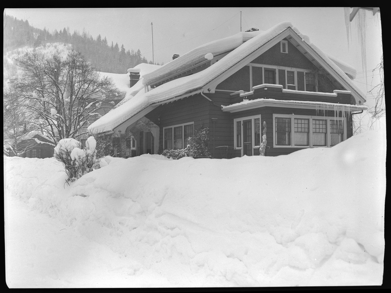Photo of a snow covered house in Wallace, Idaho. There are several inches of snow accumulated on the roof, and more so piled up on the ground in front of the house.