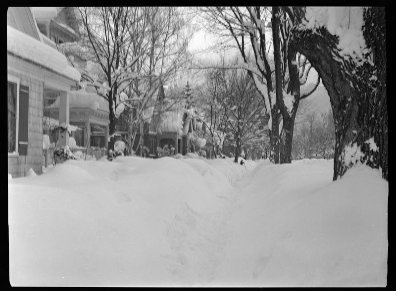 Photo looking down a residential sidewalk that shows many houses and yards covered in snow. The roofs of the houses, the trees, and the ground has several inches of snow built up, and in the distance someone appears to be shoveling. There is an indent in the snow on what is presumably the sidewalk.
