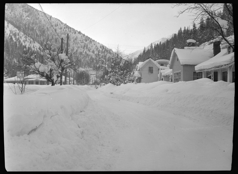 Photo of a snow covered street in Wallace, Idaho, that had been previously plowed. There are piles of snow on either side of the road, and houses behind those snow piles that have several inches of snow on the roofs.