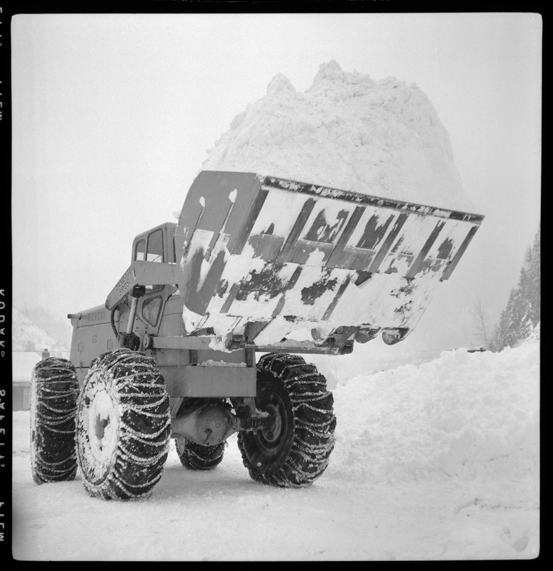 Photo of a large snow plow with the bucket of the plow overflowing with snow as the machine lifts it into the air.