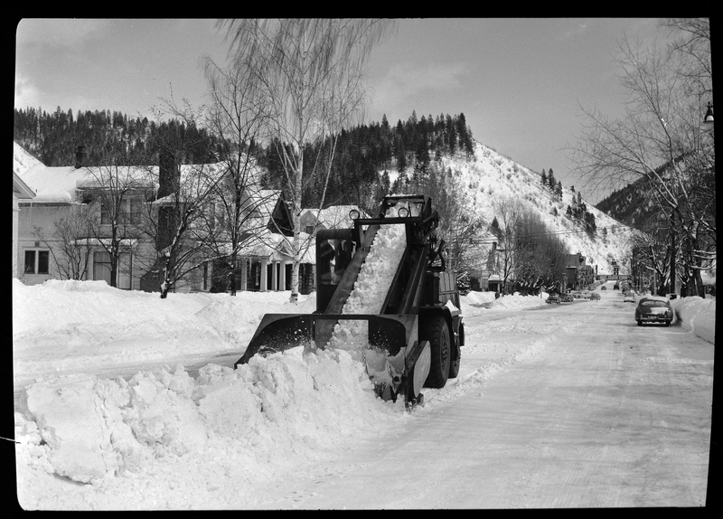 Photo of a L. C. Slisou snow plow plowing the middle of a residential street. Both sides of the road have already been plowed, leaving the middle. There are cars parked on the sides of the road where tall snow piles also exist, and there is a line of houses going down the street.