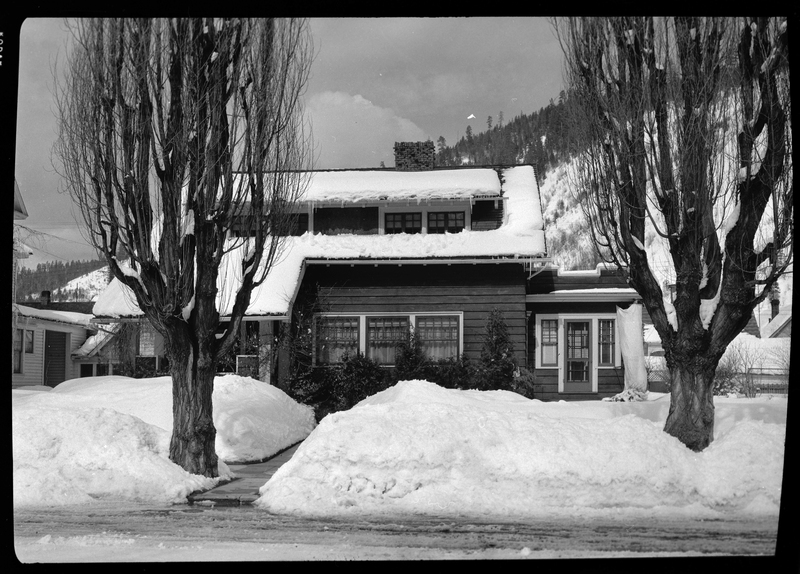Photo of a house covered in snow. The sidewalks are shoveled, but there are several tall piles of snow surrounding the cleared area. The house has snow on the roof, and the trees in front of it are also covered in snow.