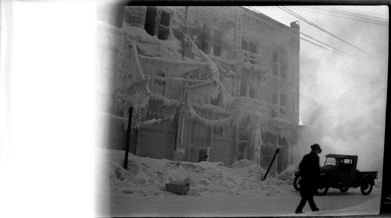 Photo of an unidentified building that is covered in icicles at Jack Waite Mine. There is a man walking on the street near the photographer, and a car is parked on the side of the road. The left part of the image is damaged and nothing is visible on that part.