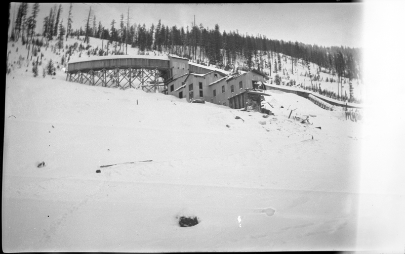 Photo of an unidentified building that is covered in snow up on a hill at Jack Waite Mine. The edge of the negative is damaged and nothing is visible there, but most of the photo is in tact. Trees are seen further up the hill behind the building.
