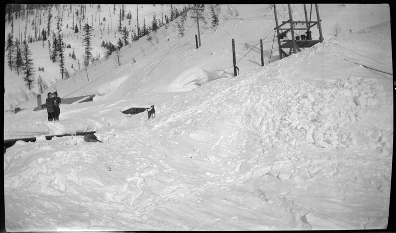 Photo of two people and two dogs standing in the snow at Jack Waite Mine. The people are standing together, one of the dogs stands off to the side, and the other dog is laying down alone in the snow. The snow comes up just below the knees of the people.