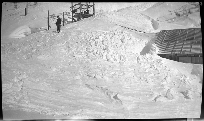 Photo of a person standing on a snow covered hill at Jack Waite Mine. There is a building off to the side and parts of the accumulated snow is taller than the building itself.