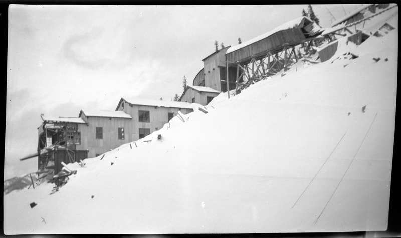 Photo of damaged buildings at Jack Waite Mine. Most of the structures stills tand, but part of either side of the connected structures are either missing, structurally unsound (tipping over but not fallen), or destroyed. There is heavy snow on the ground, but it is not clear if the snow caused the damage.