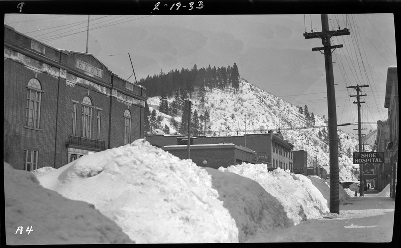 Photo of a long pile of snow in Wallace, Idaho, possibly on Pine Street or King Street. The Benevolent and Protective Order of Elks building is visible, as well as other shops down the street. The color variation in the snow is due to shadows from the surrounding structures.