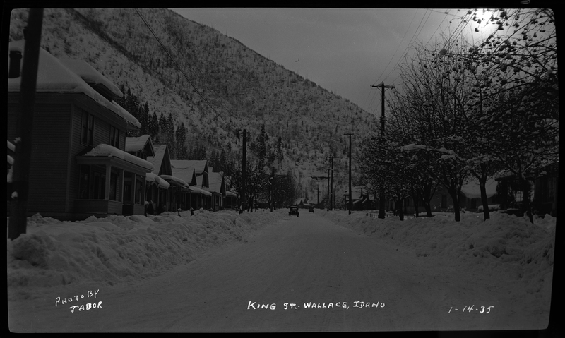 King Street in Wallace, Idaho covered in snow. There are piles of snow on either side of the road from previous plowing, and the trees and houses lining the street are also covered in snow. There are two cars visible down the road that appear to be driving.