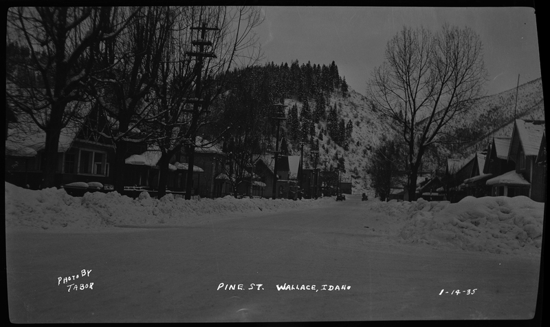 Pine Street in Wallace, Idaho covered in snow. There are piles of snow on either side of the road from previous plowing, and the trees and houses lining the street are also covered in snow. There are two cars visible down the road that appear to be driving.