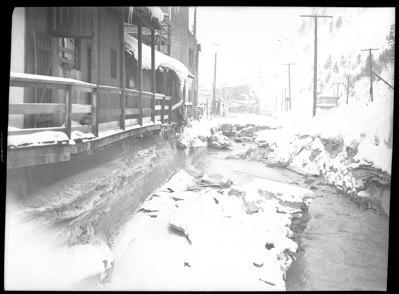 Photo of a creek running alongside some buildings in Wallace, Idaho, with snow surrounding the area. The buildings next to the creek have snow and icicles on them, as does the ground next to the creek. The water in the creek does not appear to be frozen over. Possibly from Pine Street or King Street.