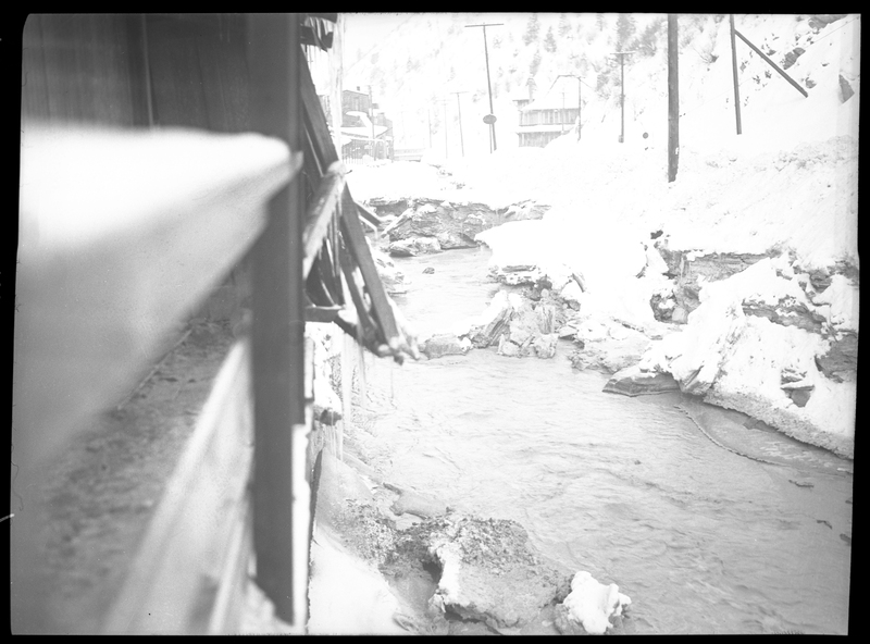 Photo of a creek running alongside some buildings in Wallace, Idaho, with snow surrounding the area. The buildings next to the creek have snow and icicles on them, as does the ground next to the creek. The water in the creek does not appear to be frozen over. Possibly from Pine Street or King Street.