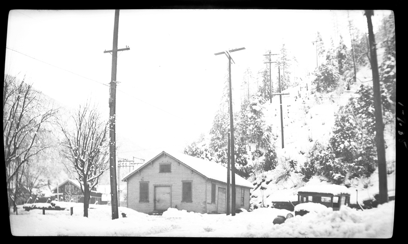 Photo of a house and a parked car in the snow in Wallace, Idaho, possibly on Pine Street or King Street. There is a hill right behind the house covered in trees and snow.