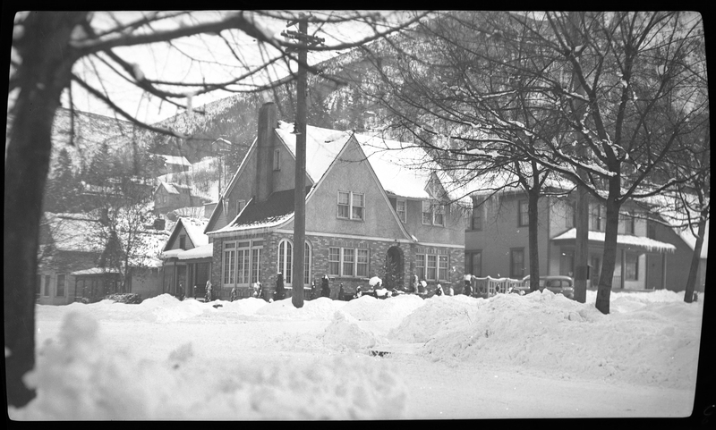 Photo of a house on the street corner in Wallace, Idaho, possibly on Pine Street or King Street. The house and those surrounding it have several inches of snow on the roofs and in the yards. There is car that has been cleared of snow parked in front of the house, and though the streets do still have snow on them, they have been plowed recently.