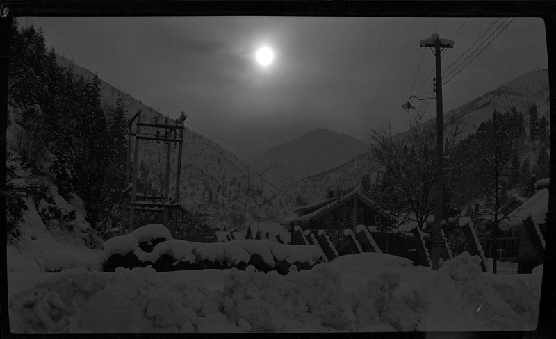 Photo looking towards the mountains behind some homes in Wallace, Idaho, possibly on Pine Street or King Street. There are piles of snow in the foreground, and snow covered houses are visible heading back towards the mountains in the background. The light from the sun makes visibly lower.