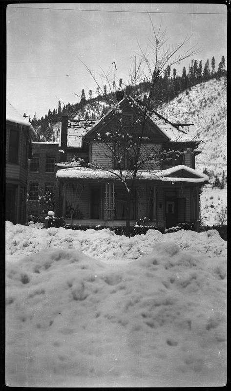 Photo of a house covered in old snow in Wallace, Idaho, possibly on Pine Street or King Street. The photo was taken across the street from the house, and the road between the house and the photographer had been plowed, leaving piles of snow on the sides. The hill and trees behind the house are covered in snow as well.