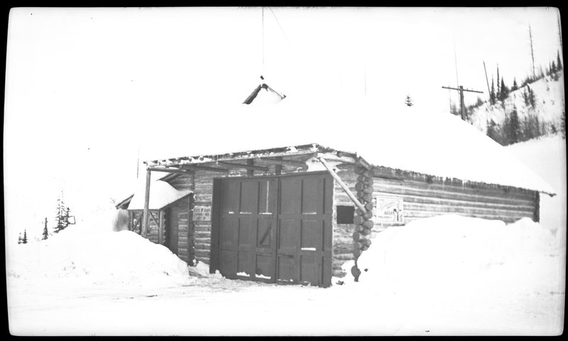 Photo of an unidentified snow covered building in Wallace, Idaho, possibly on Pine Street or King Street. The roof, which is covered with snow, has an antenna on it. The area in front of the garage had been cleared of snow, leaving piles on either side. 