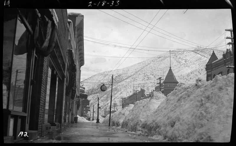 Photo of a tall, long snow pile in Wallace, Idaho, along Bank Street. The snow pile is down the middle of the road with buildings on either side of it, and appears to be several feet tall.