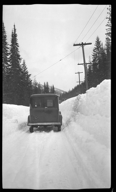 Photo of a car driving down a snowy road away from the photographer. The snow on the right side of the car is piled higher than the roof of the car, and trees can be seen in the background.
