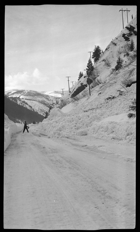Photo of an unidentified person shoveling snow off the road. There is several feet of snow on the ground in the areas not shoveled, and there are trees in the background.