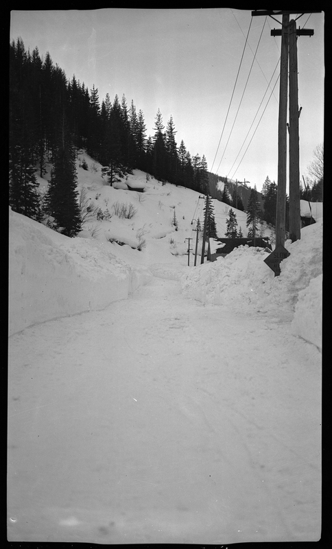 Photo of a street that has been plowed, but is still covered in a heavy layer of snow. The sides of the road are covered in several feet of snow and trees can be seen in the background.