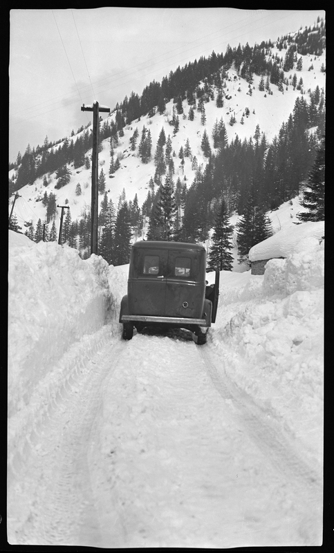 Photo of a car parked in the snow on a poorly plowed road. The road is only big enough for one car to drive down it, and snow is piled up as high as the car's roof on either side. The car is stopped and the passenger door is open.