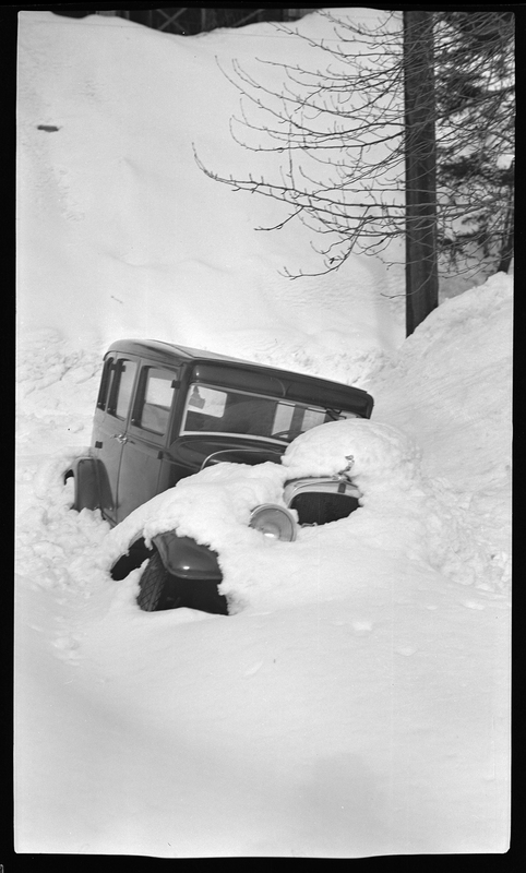 Photo of a car that is buried in snow. The snow comes up above the roof on one side, up to the doors on the other side. There is freshly fallen snow on the hood and covering most of the front.