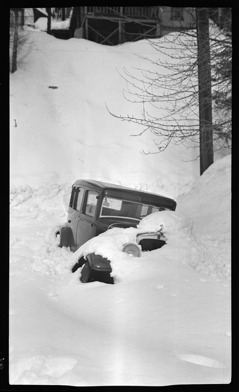Photo of a car that is buried in snow. The snow comes up above the roof on one side, up to the doors on the other side. There is freshly fallen snow on the hood and covering most of the front.