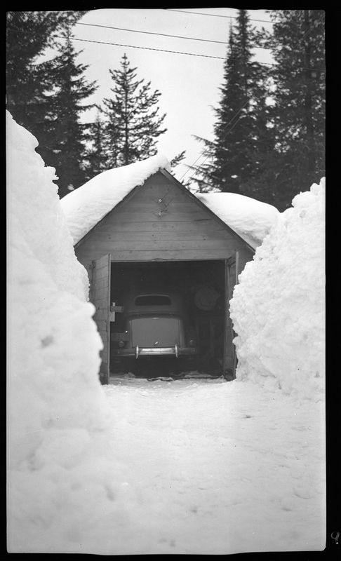 Photo of a car parked inside of a garage that has the doors open, surrounded by heavy snow. The snow is piled up over the roof of the garage, which is also covered in snow. The ground between the photographer and the car appears to be shoveled.