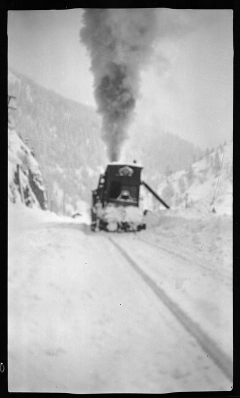 Blurry, unfocused photo of a train moving through heady snow. The engine is visible and moving towards the photographer, with smoke emitting from the top and snow covering the front. The snow on the other side of the train tracks is piled high.