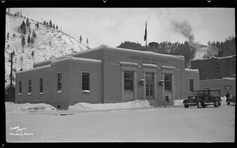 Photo of the United States Post Office building in Wallace, Idaho, in the snow. There is a car parked out in front of the building.