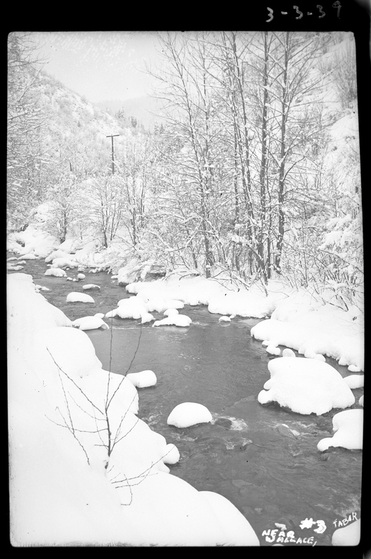 Photo of a creek in Wallace, Idaho that is surrounded by snow. The water does not appear to be frozen over, but the rocks that are above the water are covered in snow, as is the ground next to the creek and the trees.