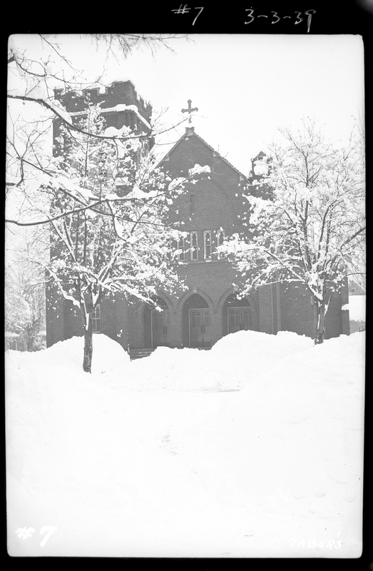 Photo of the snow covered Catholic Church in Wallace, Idaho. Snow covered trees surround the building, and the ground around it is also covered in snow. There are large piles of snow in front of the doors to the building.