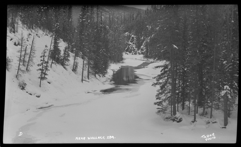 Photo of a snow scene in a wooded area near Wallace, Idaho. The ground and surrounding trees are covered in trees. There is a body of water that appears to be partially frozen over.