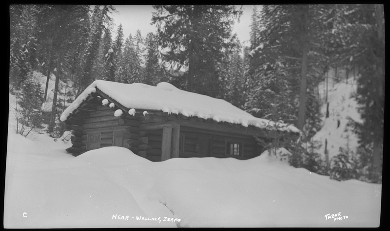 Cabin in the woods surrounded by and covered in snow near Wallace, Idaho. The snow is piled up high and comes up halfway up the cabin.