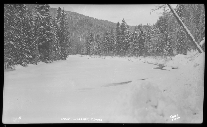 Photo of a snow scene in a wooded area near Wallace, Idaho. The ground and surrounding trees are covered in trees. There is a body of water that appears to be partially frozen over.