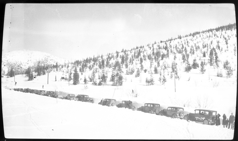 Photo of a line of cars parked on the road near Lookout Pass on the Wallace-Missoula Highway. There are people standing around the cars and trees on the snow covered ground in the background. Previously described as "Near Steven's Peak."