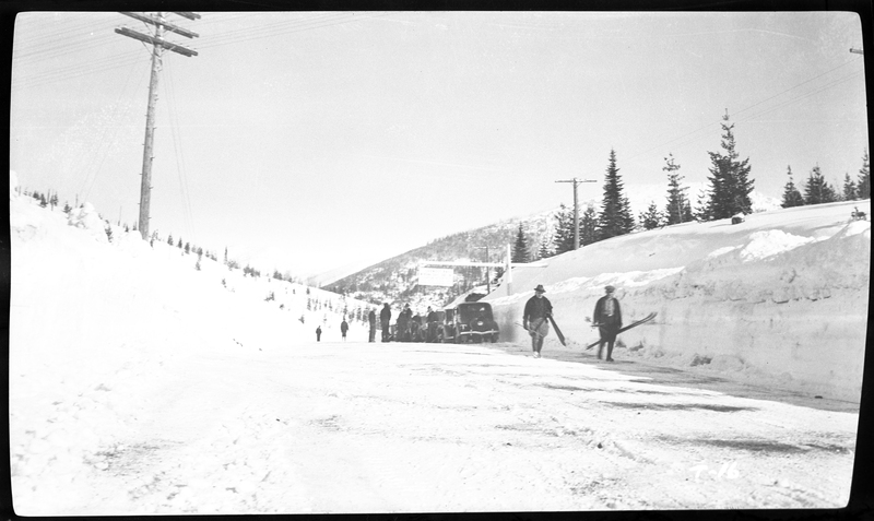 A line of cars is parked on the side of a snowy road and a large group of people are gathered near them near Lookout Pass on the Wallace-Missoula highway. Two unidentified people walk towards the photographer and they are carrying ski equipment. Previously described as "Near Steven's Peak."