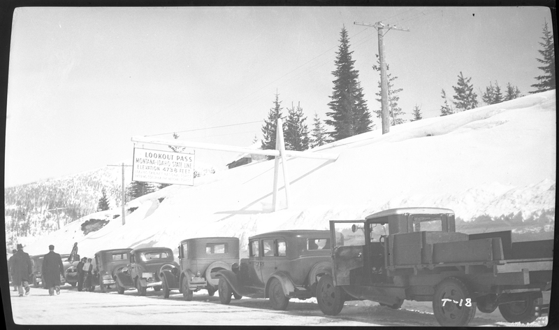 Photo of a line of cars parked on the road near Lookout Pass on the Wallace-Missoula Highway. There are people standing around the cars and trees on the snow covered ground in the background. A sign hangs over the cars that reads, "Lookout Pass; Montana-Idaho State Line; Elevation 4738 Feet." Previously described as "Near Steven's Peak."