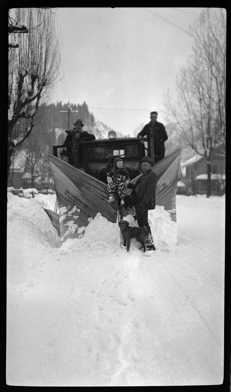 Three men, a child, and a dog stand with a snowplow. Two of the men stand on the snow plow while the third man, the child, and the dog are in front of it. The child is sitting on top of the scoop for the plow, while the man and the dog are standing.