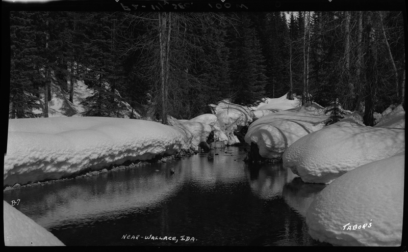Photo of a snowy river near Wallace, Idaho. The ground surrounding the water is covered in a heavy layer of snow, but the trees visible in the background are not.