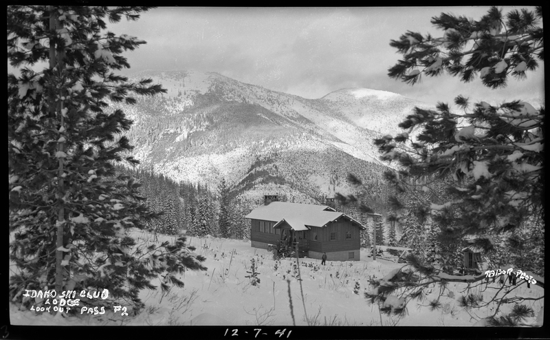 Photo of the Idaho Ski Club Lodge at Lookout Pass. The building is in the distance in an open area from the trees, and the photographer is standing back at the tree line. There is a person walking in front of the lodge. Mountains can be seen behind it with trees covering them.