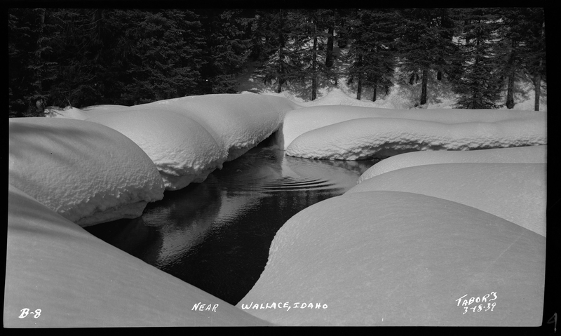 Photo of a small body of water, likely a pond, with heavy snow covering the ground surrounding it. There are trees in the background.