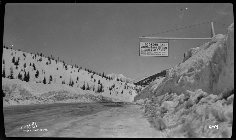 Photo of the Lookout Pass sign on the Wallace-Missoula Highway. Sign reads, "Lookout Pass; Montana-Idaho State Line; Elevation 4738 Feet; Leaving Cabinet National Forest; Entering Coeur d'Alene National Forest." There is heavy snow on the ground and trees in the background.