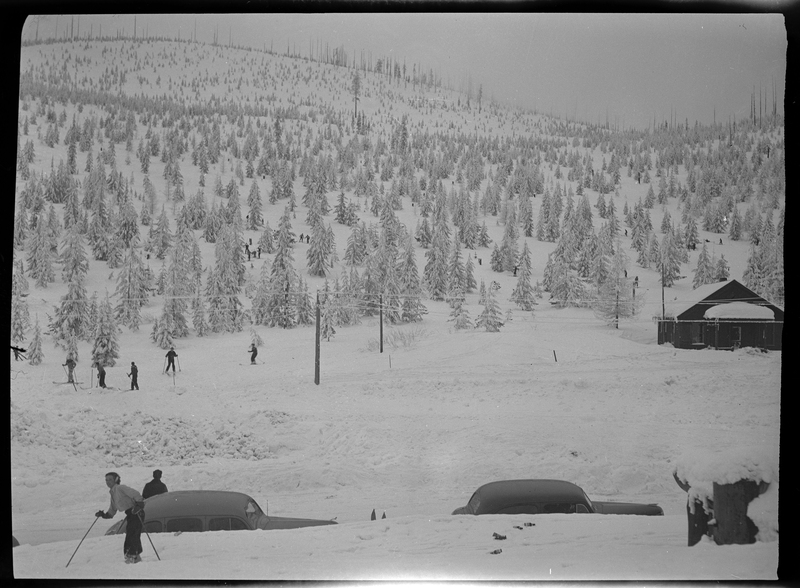 Photo of skiers in the snow at Lookout Pass. A few people are in the foreground, but most are in the background. There are two cars parked on the side of the road.