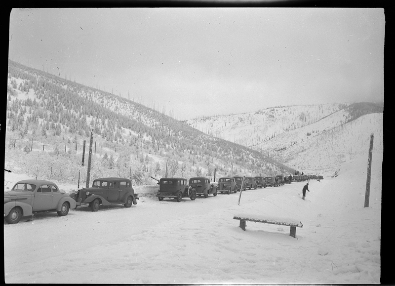 Photo of a line of cars parked on the side of the road in the snow at Lookout Pass. An unidentified person can be seen skiing away from the cars. There is a single snow covered bench in front of the photographer.