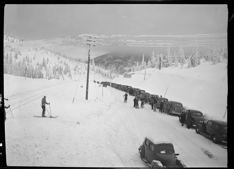 A line of cars are parked on the side of a snowy road at Lookout Pass. There is a group of people wearing ski gear standing next to the cars.